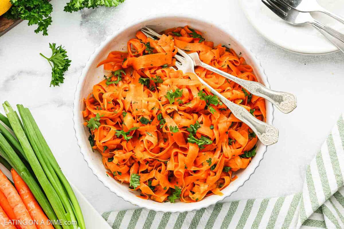 A bowl of Carrot Salad with ribboned carrots is garnished with fresh herbs, two forks resting inside. Fresh parsley, green onions, and whole carrots are arranged nearby on a white surface.