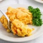 A serving of cheesy corn casserole with a golden crust on a white plate, garnished with a sprig of parsley. A spoon is lifting a portion, revealing melty cheese and corn kernels inside.