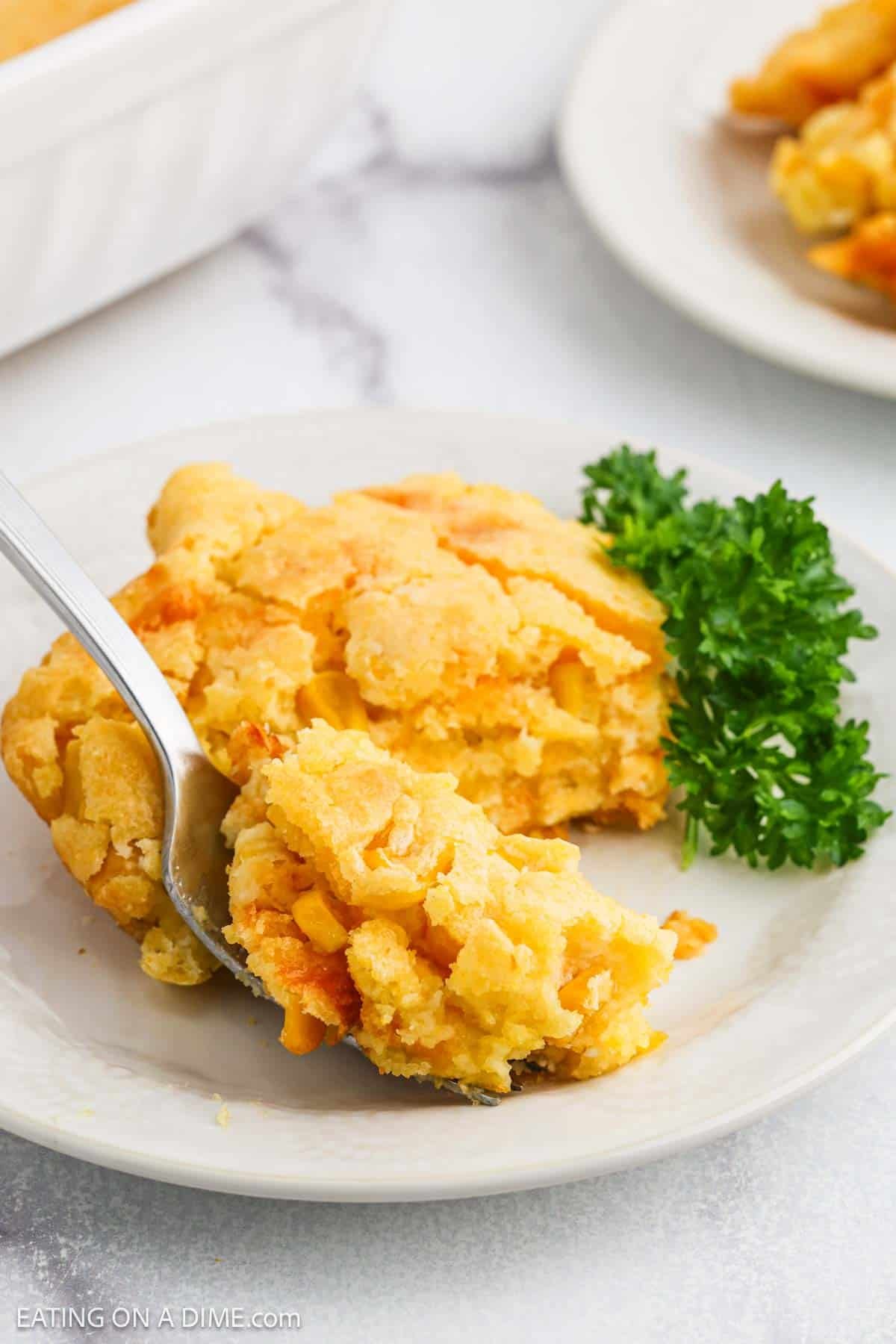 A serving of Cheesy Corn Casserole on a white plate with a fork, garnished with a sprig of parsley.