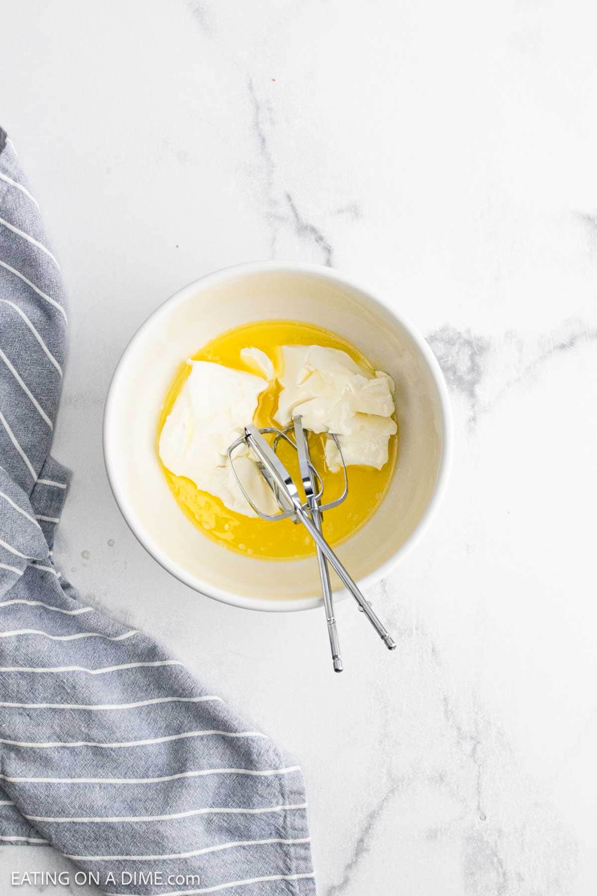 A white bowl with softened cream cheese and melted butter—perfect for making Cheesy Corn Casserole—rests on a marble surface next to a gray striped towel. Two metal beaters are inside the bowl.