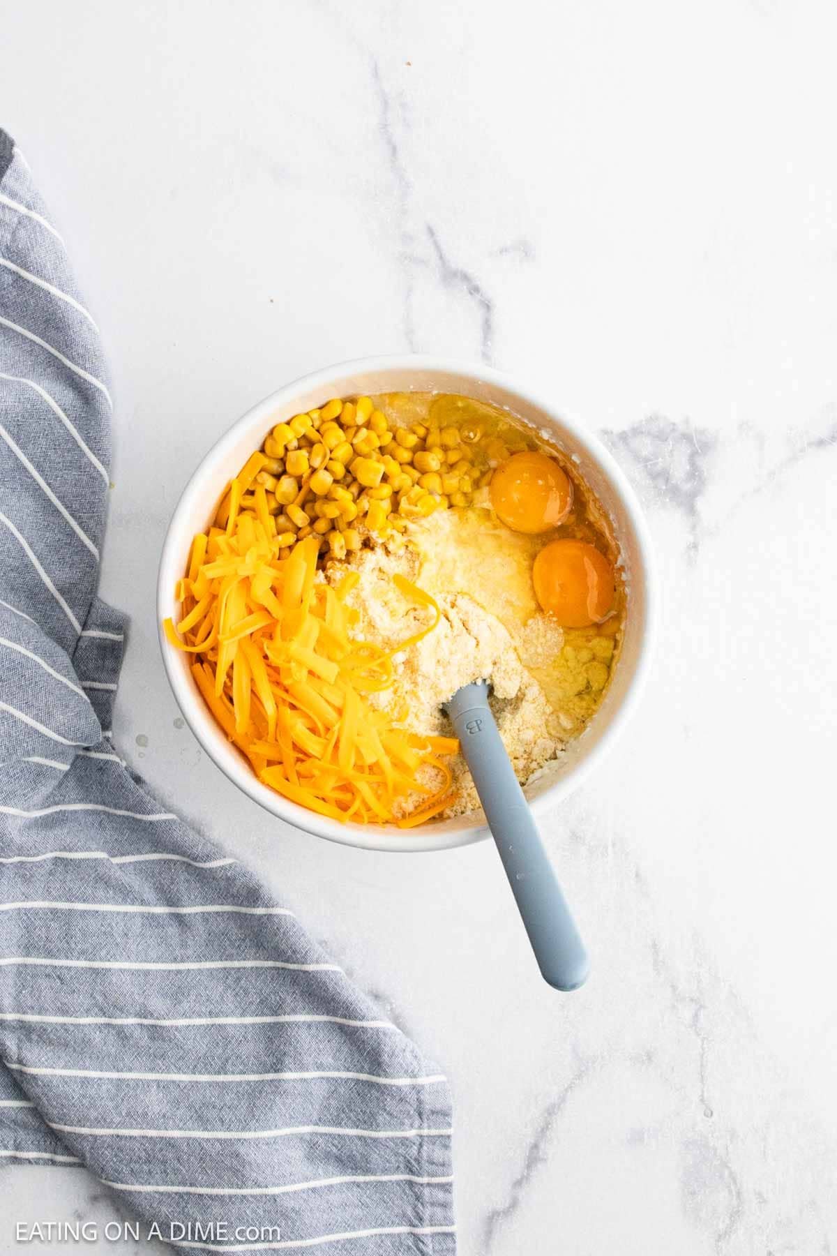 A mixing bowl with eggs, flour, shredded cheddar cheese, and corn—perfect for a Cheesy Corn Casserole—sits on a white marble surface with a blue spatula inside, next to a gray and white striped cloth.