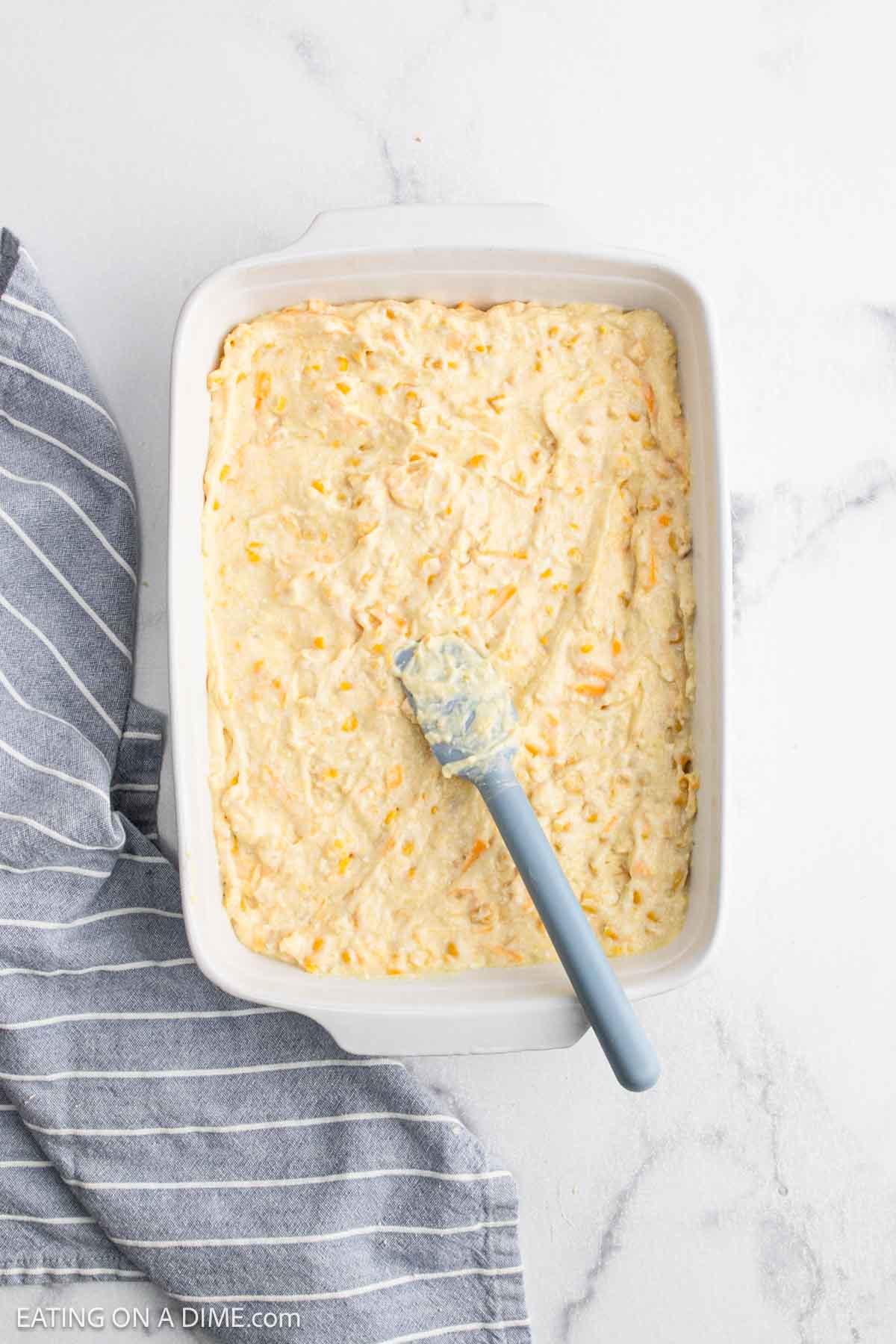 A baking dish filled with an unbaked cheesy corn casserole mixture. A blue spatula rests on top, while a gray and white striped cloth lies beside the dish on a white marble surface.
