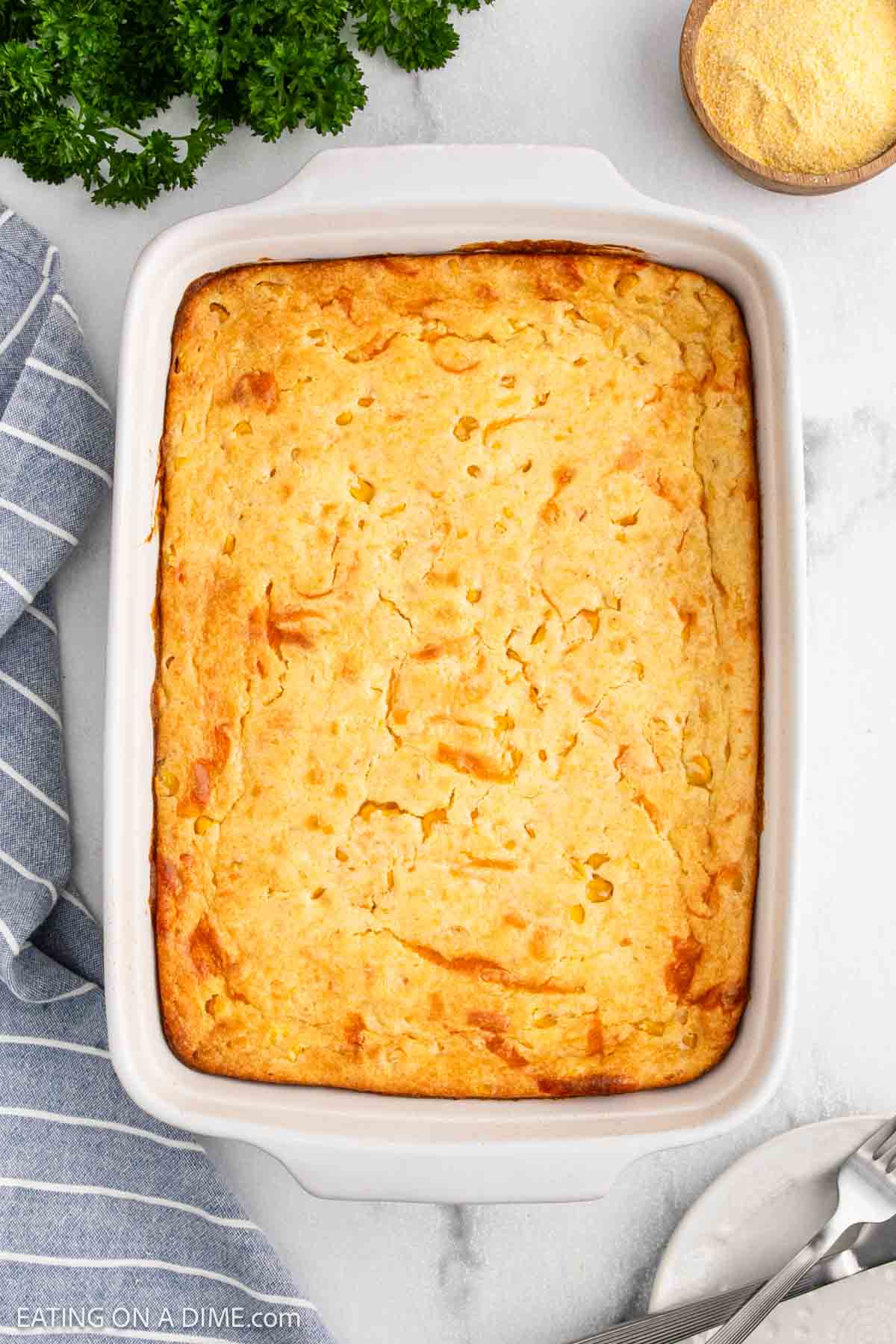 A golden-brown Cheesy Corn Casserole in a white rectangular dish, set on a marble surface with a striped kitchen towel, fresh parsley, cornmeal, and silver serving utensils nearby.