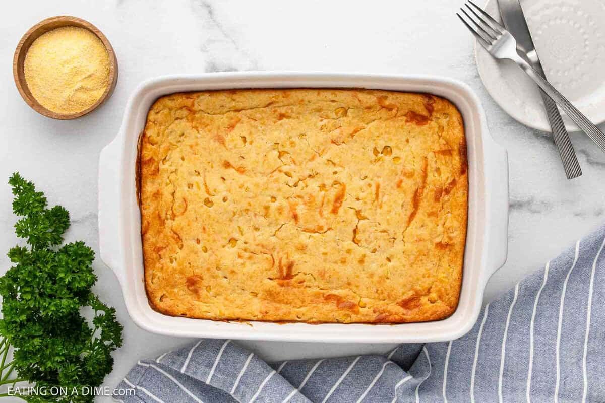 A golden-brown cheesy corn casserole in a white baking dish sits on a marble surface, surrounded by a bowl of cornmeal, fresh parsley, forks, a plate, and a blue striped towel.