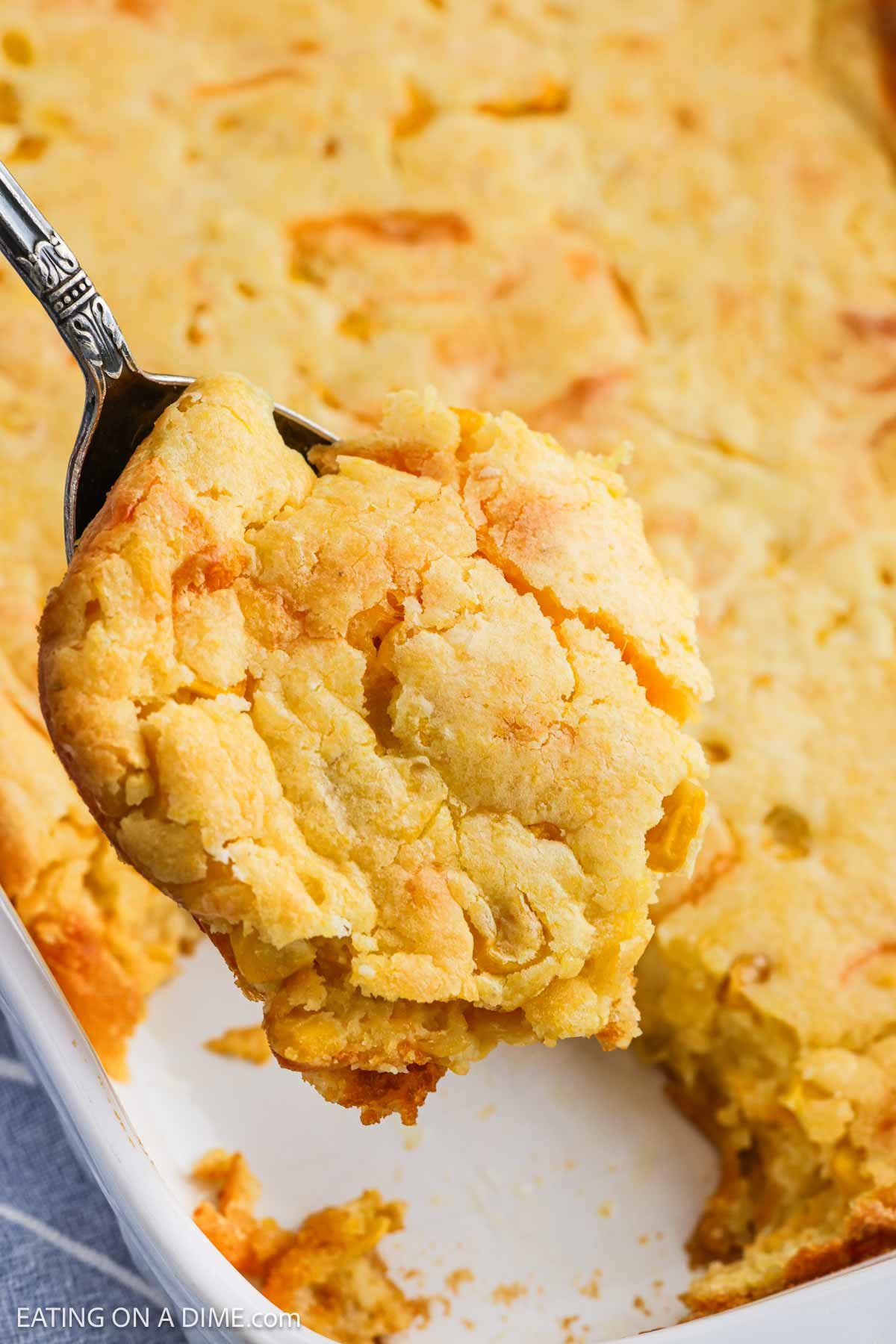 A close-up of a spoon lifting a serving of golden, crumbly cheesy corn casserole from a baking dish. The casserole has a slightly crispy top and a moist, textured interior.