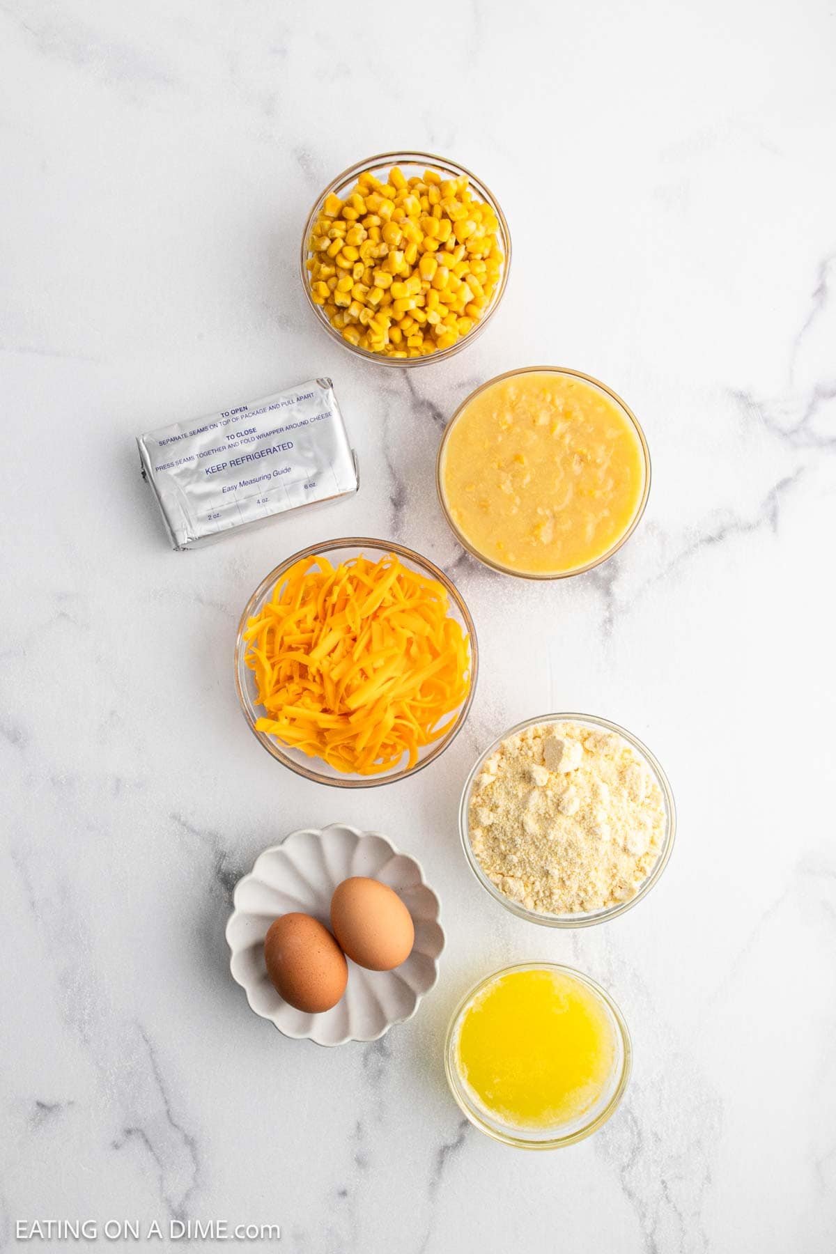 Overhead view of ingredients for cheesy corn casserole on a white surface: a can of corn, a block of cream cheese, shredded cheddar, creamed corn, two eggs, melted butter, and corn muffin mix.