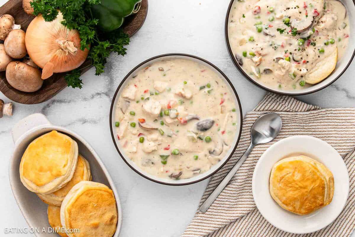 Two bowls of creamy Chicken a la King stew with mushrooms, peas, and carrots sit beside a plate of golden biscuits, a spoon, and fresh ingredients like onion, mushrooms, and parsley on a white surface.
