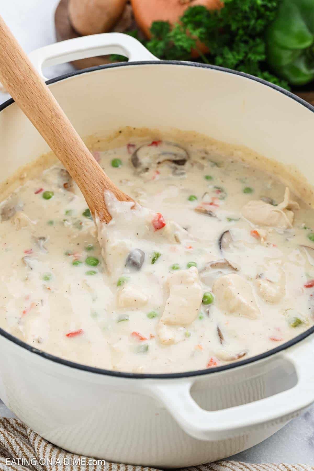 A creamy Chicken a la King stew with peas, mushrooms, and red peppers is being stirred with a wooden spoon in a white Dutch oven.