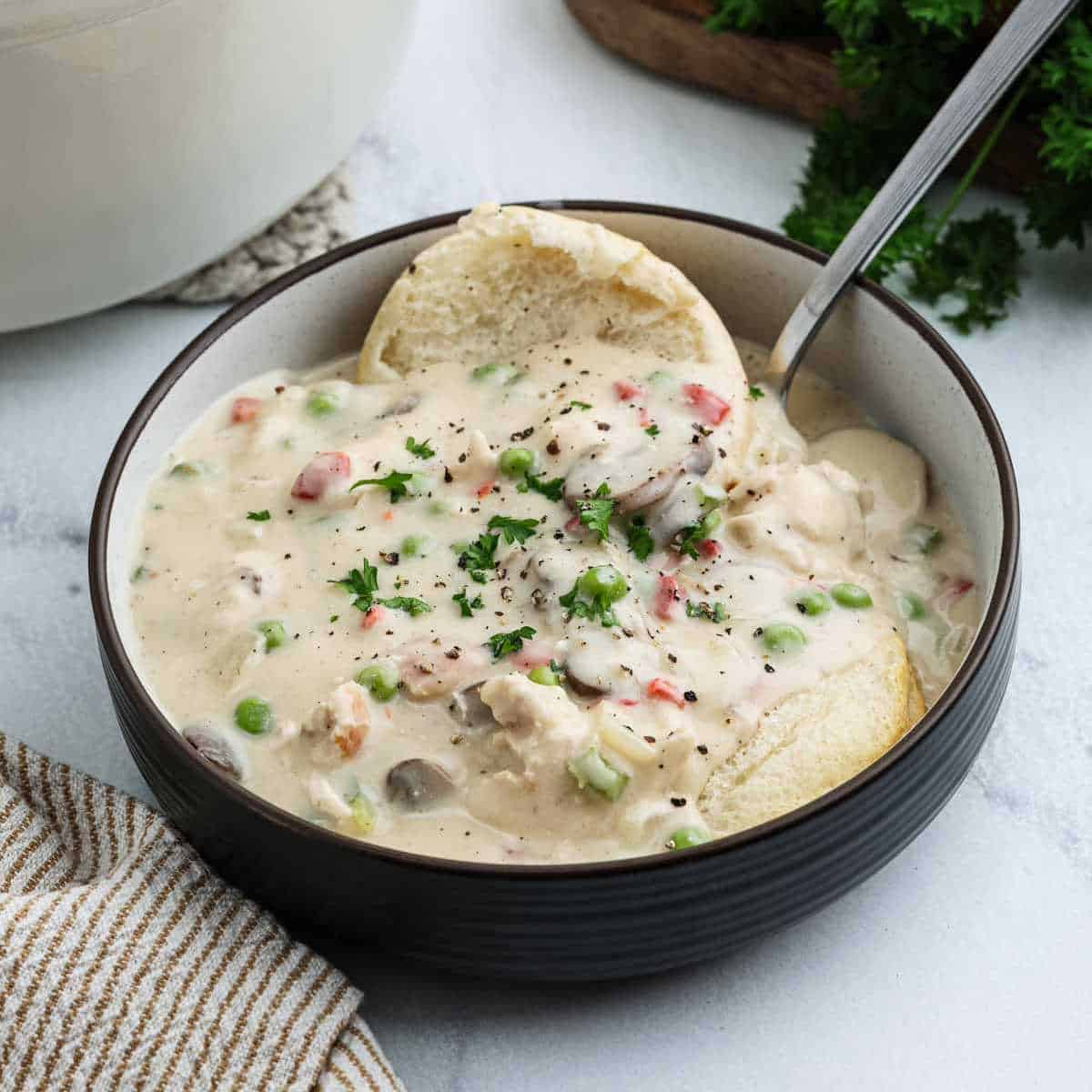 A bowl of creamy Chicken a la King-inspired pot pie soup with vegetables, garnished with herbs and black pepper, served with a biscuit and a spoon.