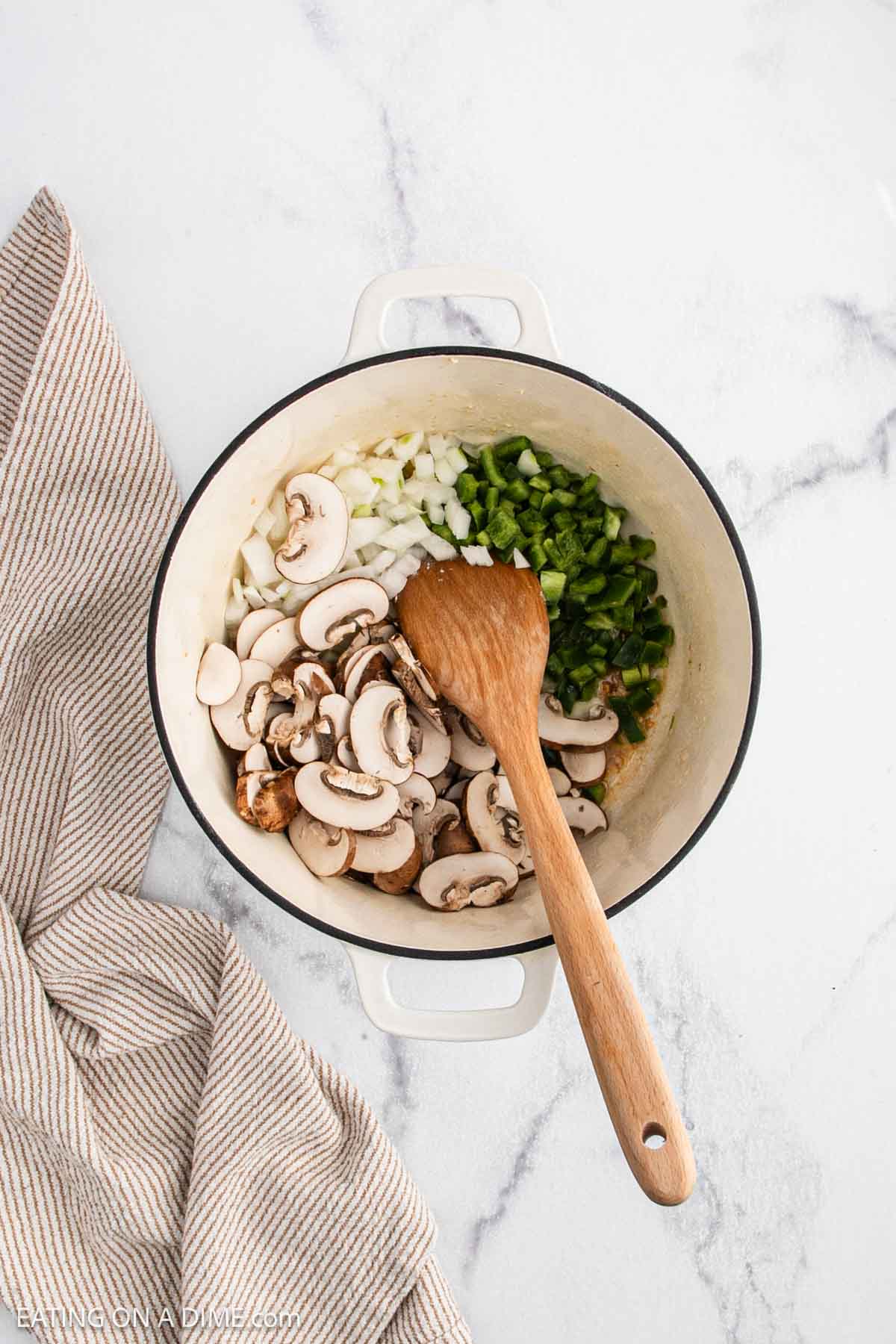 A white pot containing chopped onions, green bell peppers, and sliced mushroomsโclassic ingredients for Chicken a la Kingโis being stirred with a wooden spoon, next to a beige striped kitchen towel on a marble surface.
