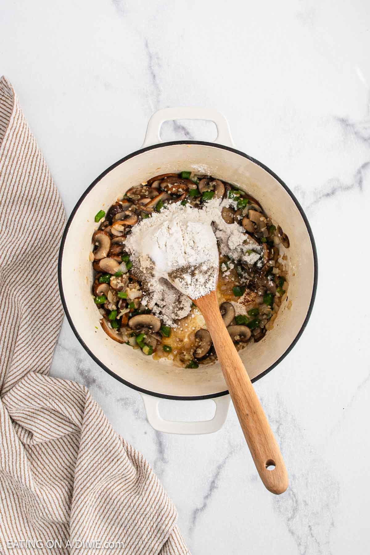A white pot on a marble surface contains sautรฉed mushrooms and green vegetables being mixed with flour for Chicken a la King using a wooden spoon. A beige striped towel lies beside the pot.