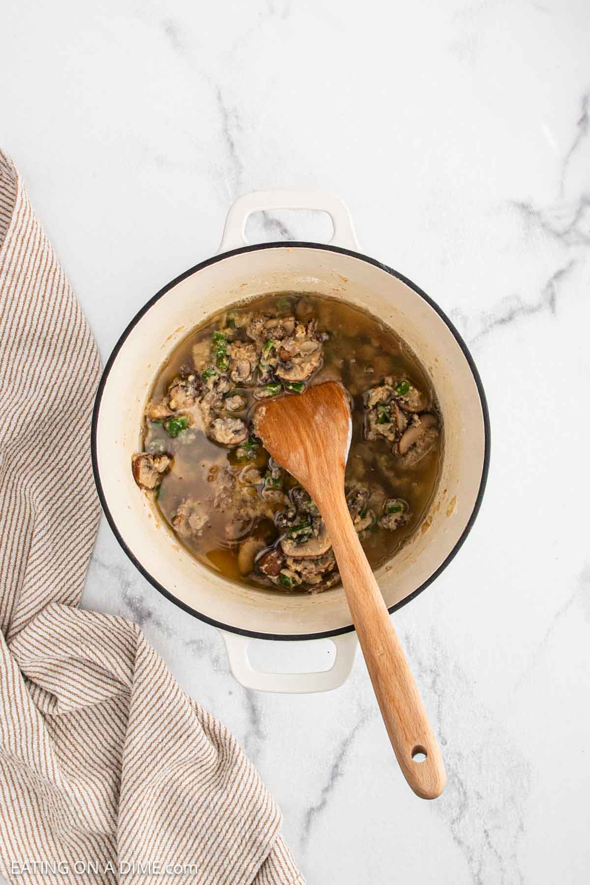 A white pot filled with Chicken a la Kingโcooked ground meat, herbs, and sauceโsits on a marble surface. A wooden spoon rests inside the pot, while a beige striped kitchen towel is draped nearby.