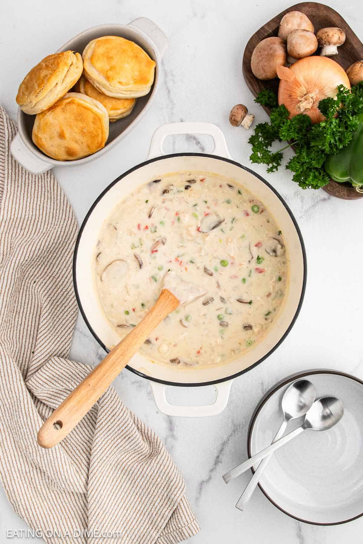 A pot of creamy vegetable soup with peas, carrots, and mushrooms reminiscent of Chicken a la King sits on a white countertop. Nearby are a plate of biscuits, fresh vegetables and parsley, a striped towel, and two stacked bowls with spoons.
