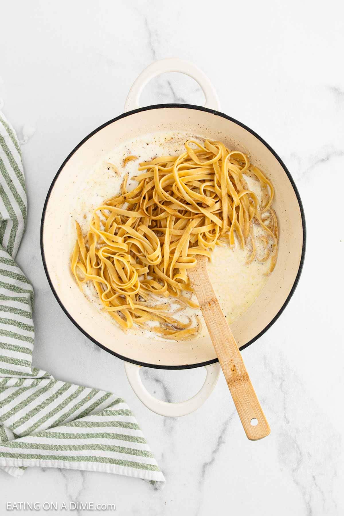 A white pot with Fettuccine Alfredo pasta being mixed in a creamy sauce using a wooden spoon. A green and white striped towel rests beside the pot on a marble surface.