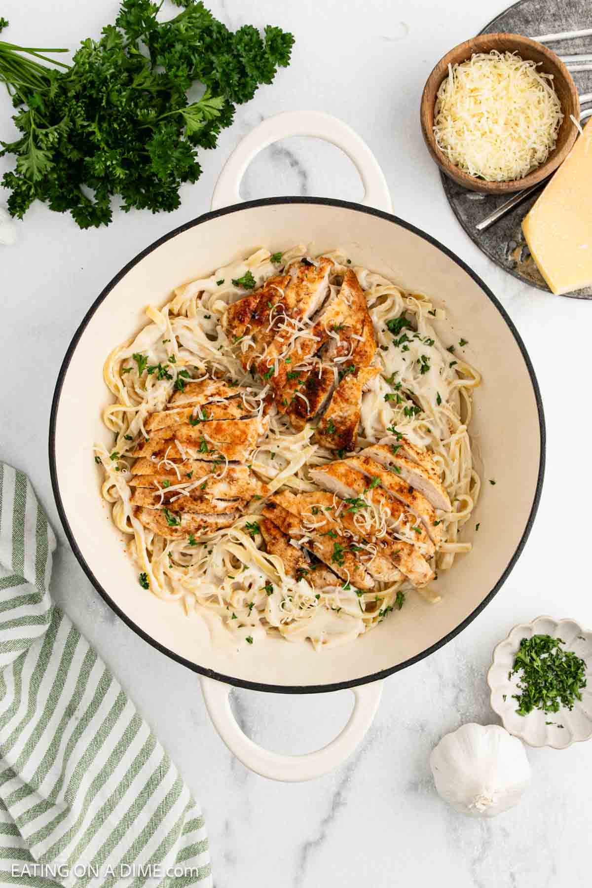 A white pot filled with Chicken Fettuccine Alfredo—fettuccine pasta and grilled chicken, garnished with chopped parsley and grated cheese. Surrounding the pot are parsley, a bowl of shredded cheese, a wedge of cheese, garlic, and a striped towel.