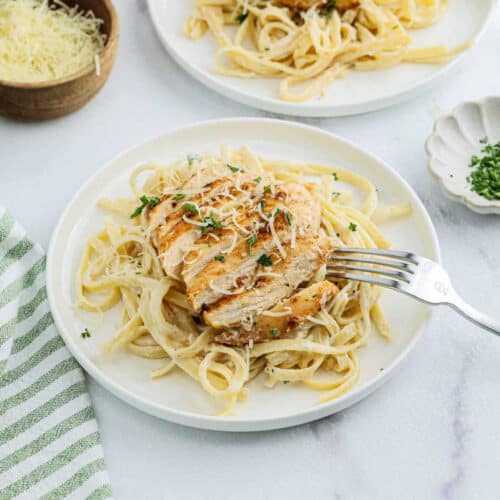 A plate of Chicken Fettuccine Alfredo topped with sliced grilled chicken breast, sprinkled with grated cheese and fresh herbs. A fork rests on the plate, with a bowl of shredded cheese and a dish of herbs nearby.