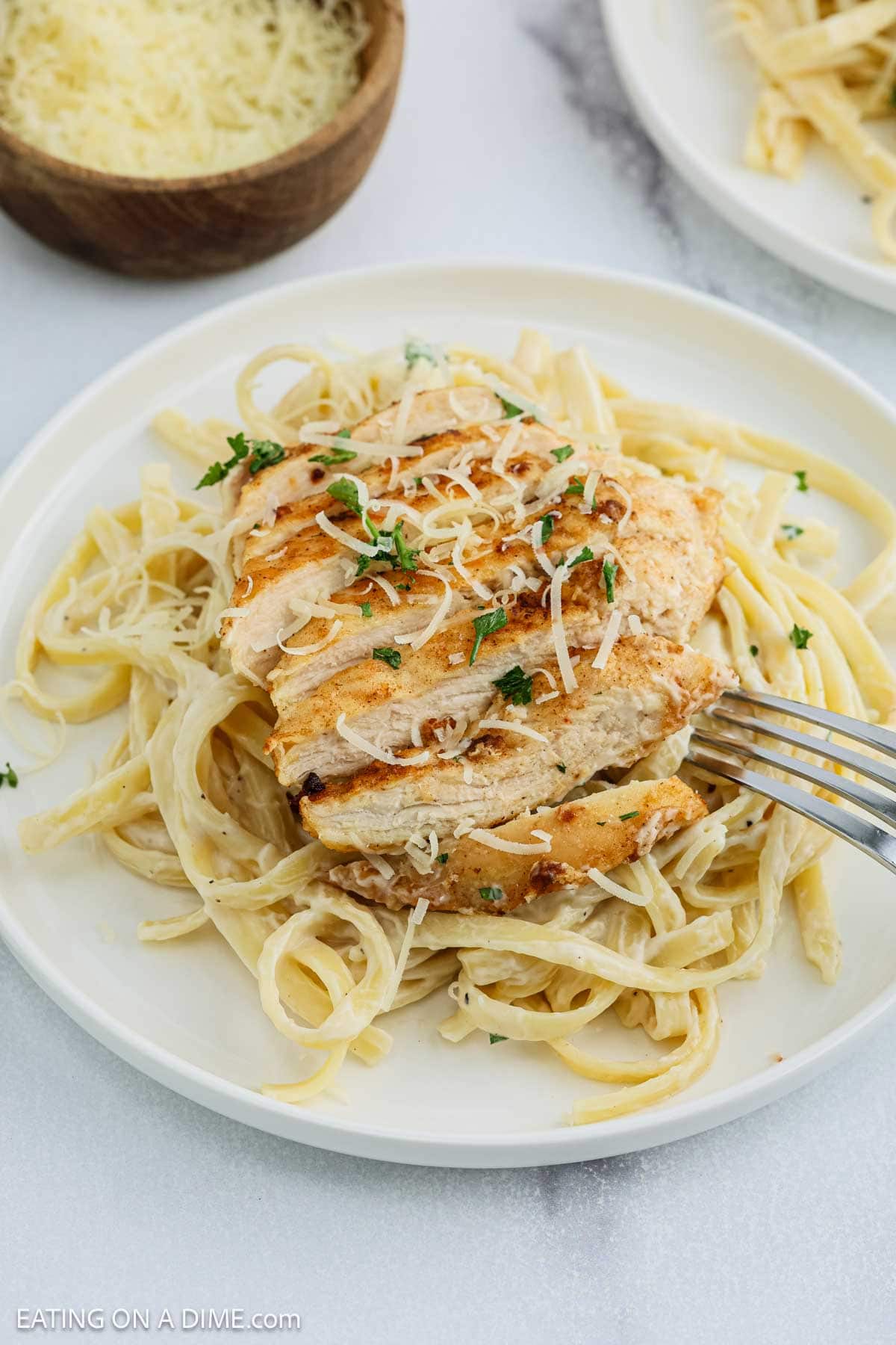 A plate of Chicken Fettuccine Alfredo topped with sliced grilled chicken breast, garnished with grated cheese and chopped parsley. A fork cuts into the chicken, while a bowl of shredded cheese sits in the background.
