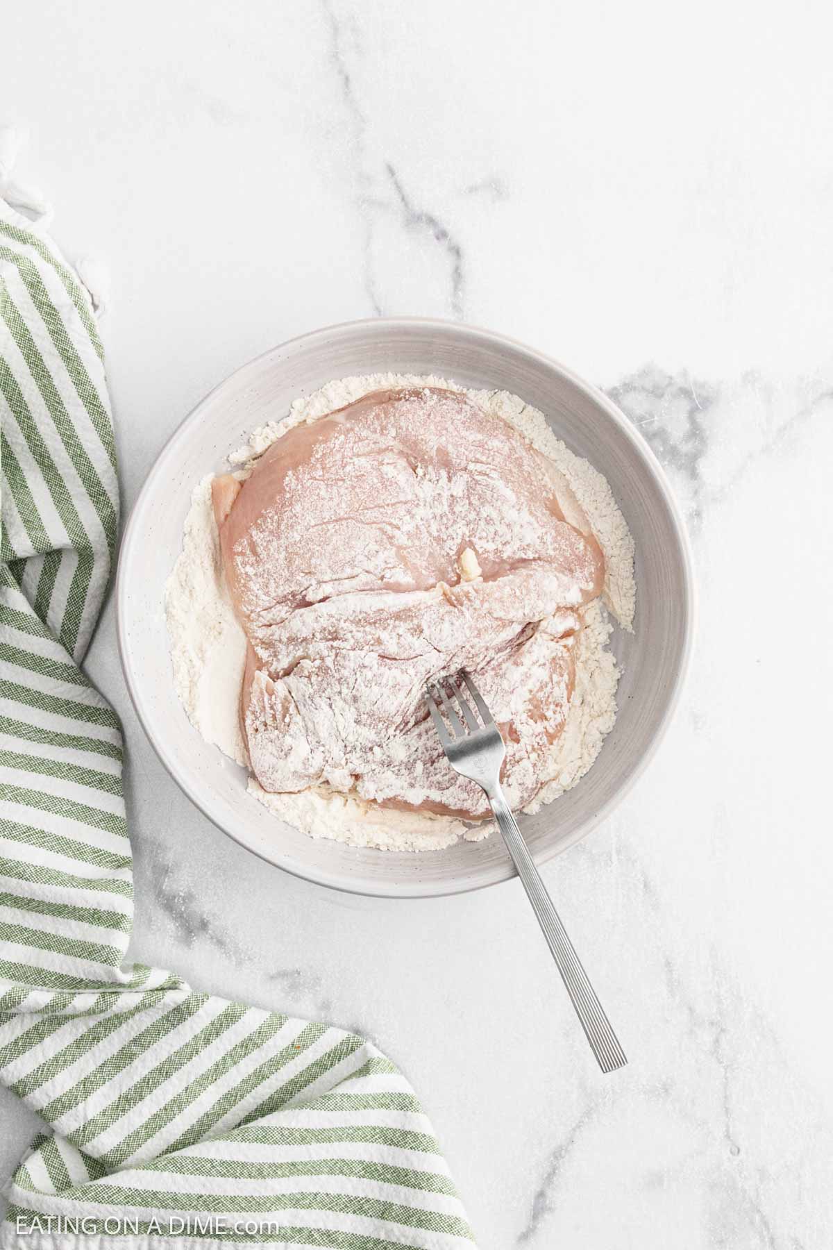 Raw chicken breast coated with flour in a white bowl, ready to be cooked for Chicken Fettuccine Alfredo. A fork rests on top, and a green and white striped kitchen towel lies beside the bowl on a marble countertop.
