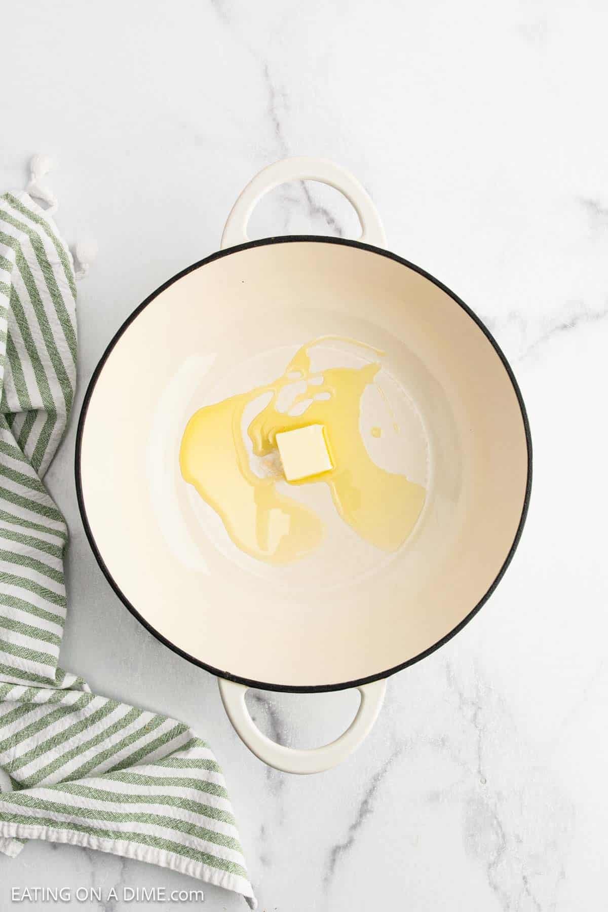 A white Dutch oven with a pat of butter and a drizzle of oil inside sits on a white marble surface next to a green and white striped kitchen towel, ready for making creamy Chicken Fettuccine Alfredo.