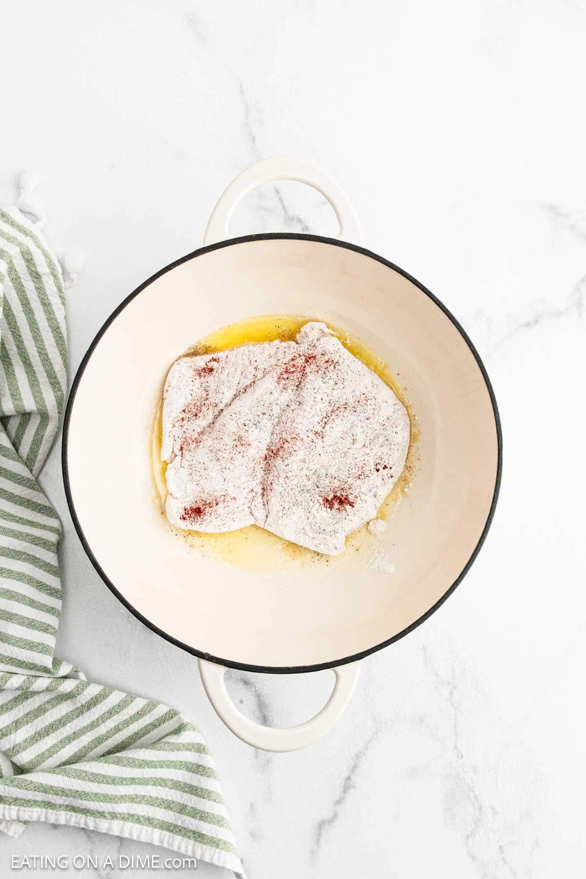 A raw, flour-coated piece of chicken with spices sits in a round white Dutch oven with melted butter, ready to be transformed into creamy Chicken Alfredo. A green and white striped towel rests on the left, atop a white marble surface.