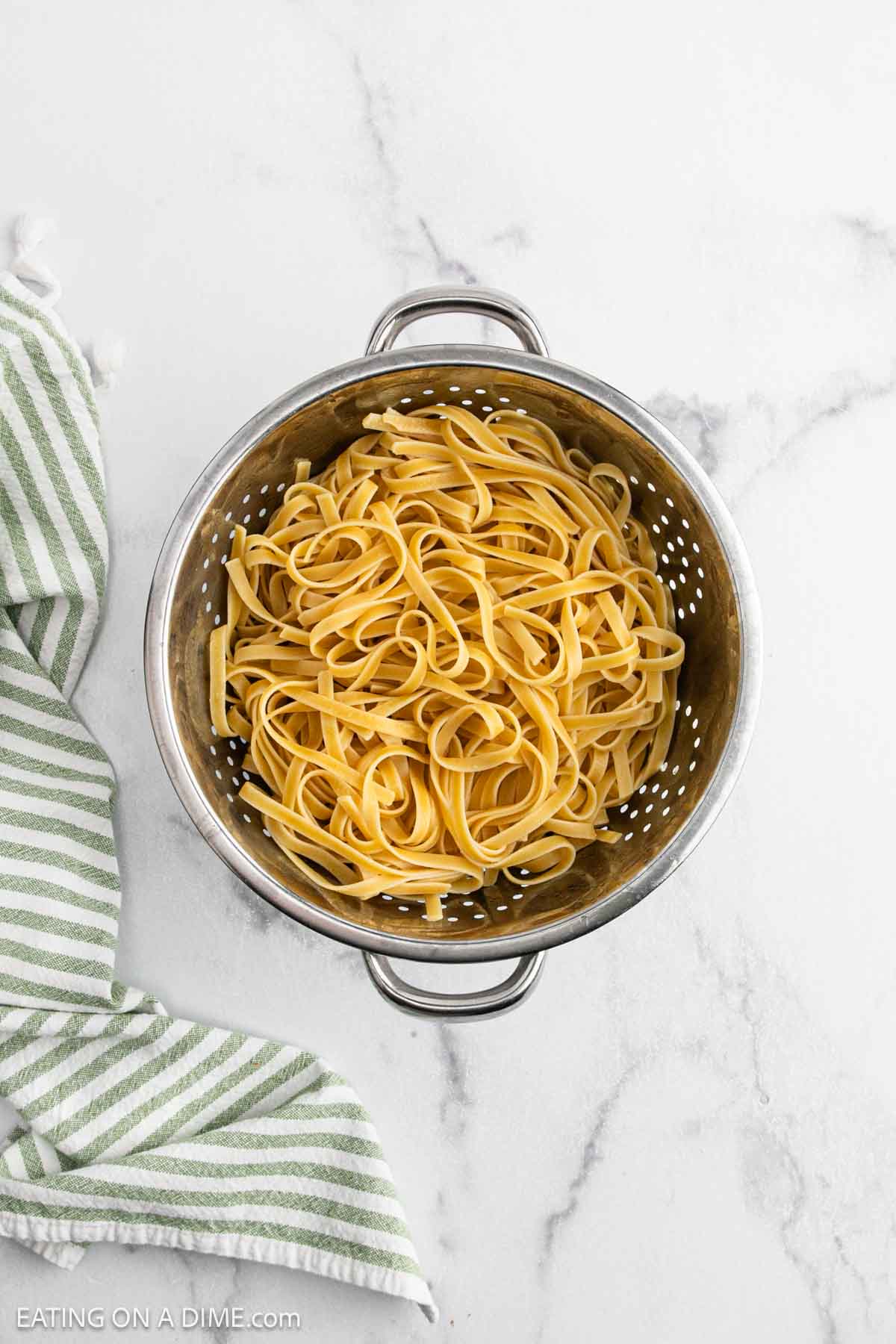 A metal colander filled with cooked fettuccine pasta, perfect for preparing classic Fettuccine Alfredo, sits on a white marble surface next to a green and white striped towel.