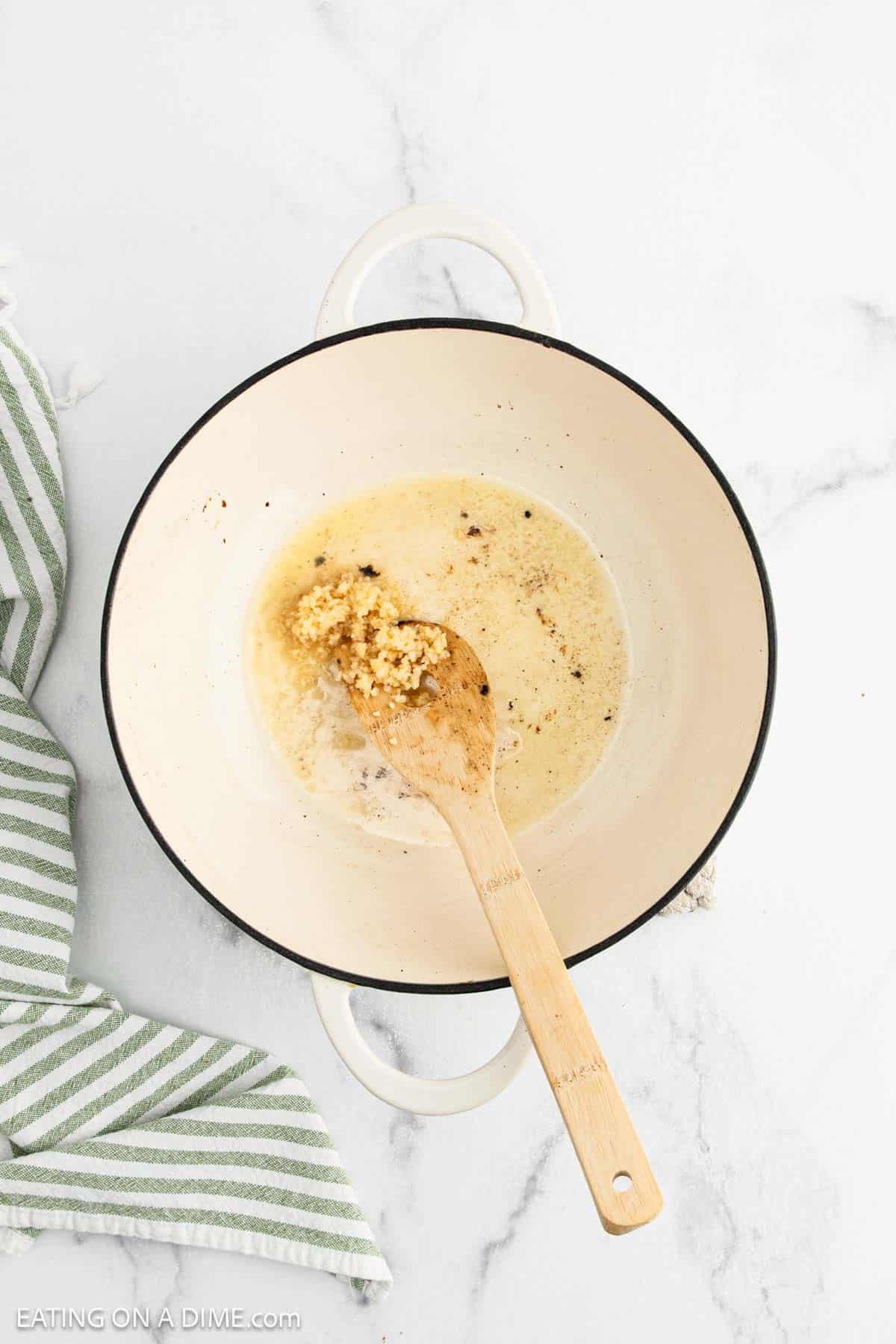 A white pot with melted butter and minced garlic being stirred by a wooden spoon, perfect as the first step in making creamy Fettuccine Alfredo, sits on a white marble surface next to a green and white striped towel.