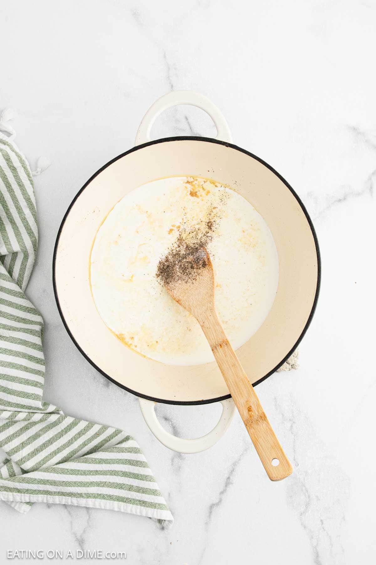 White pot with creamy sauce and spices, perfect for Chicken Fettuccine Alfredo, with a wooden spoon inside. It sits on a white marble surface next to a green and white striped towel.