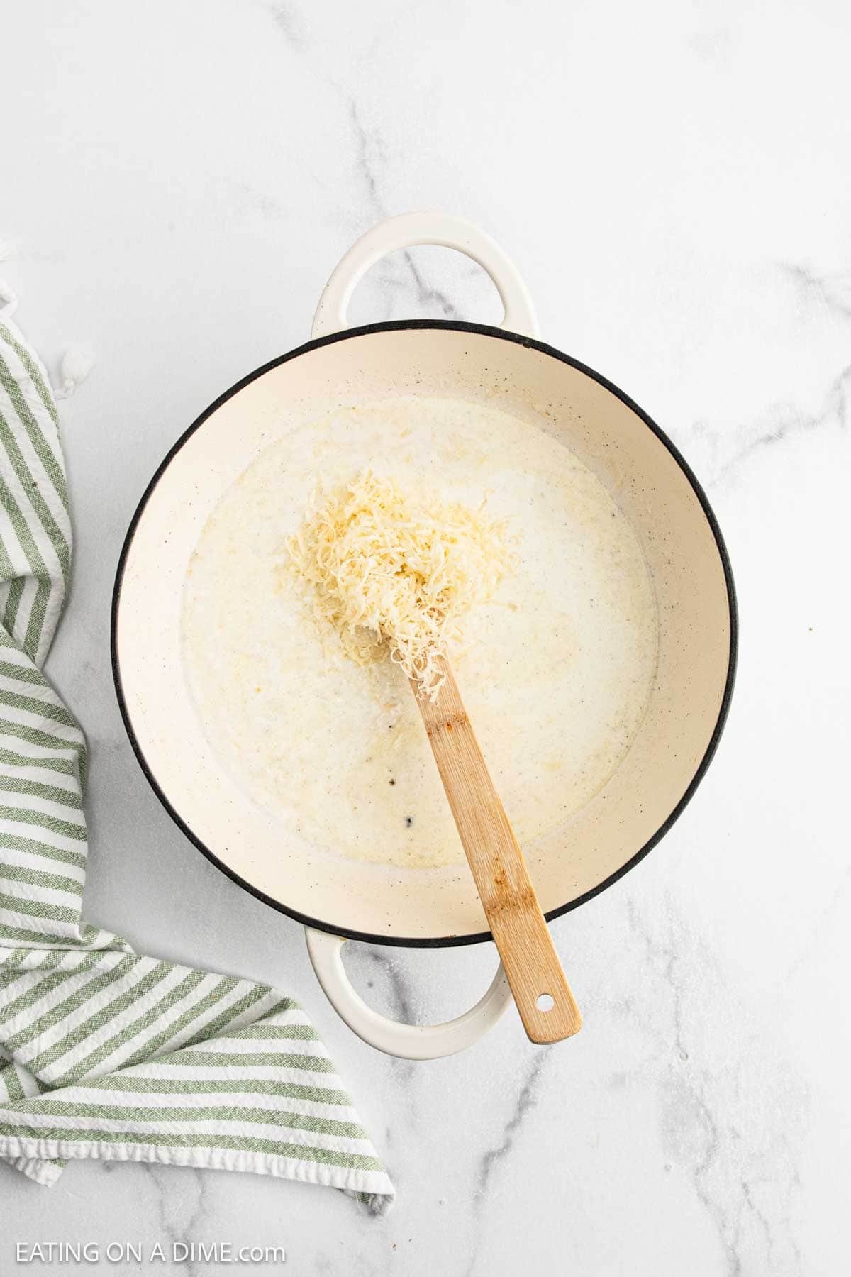 A white pot filled with creamy Chicken Fettuccine Alfredo sauce and shredded cheese is being stirred with a wooden spoon, sitting on a white marble surface next to a green and white striped towel.