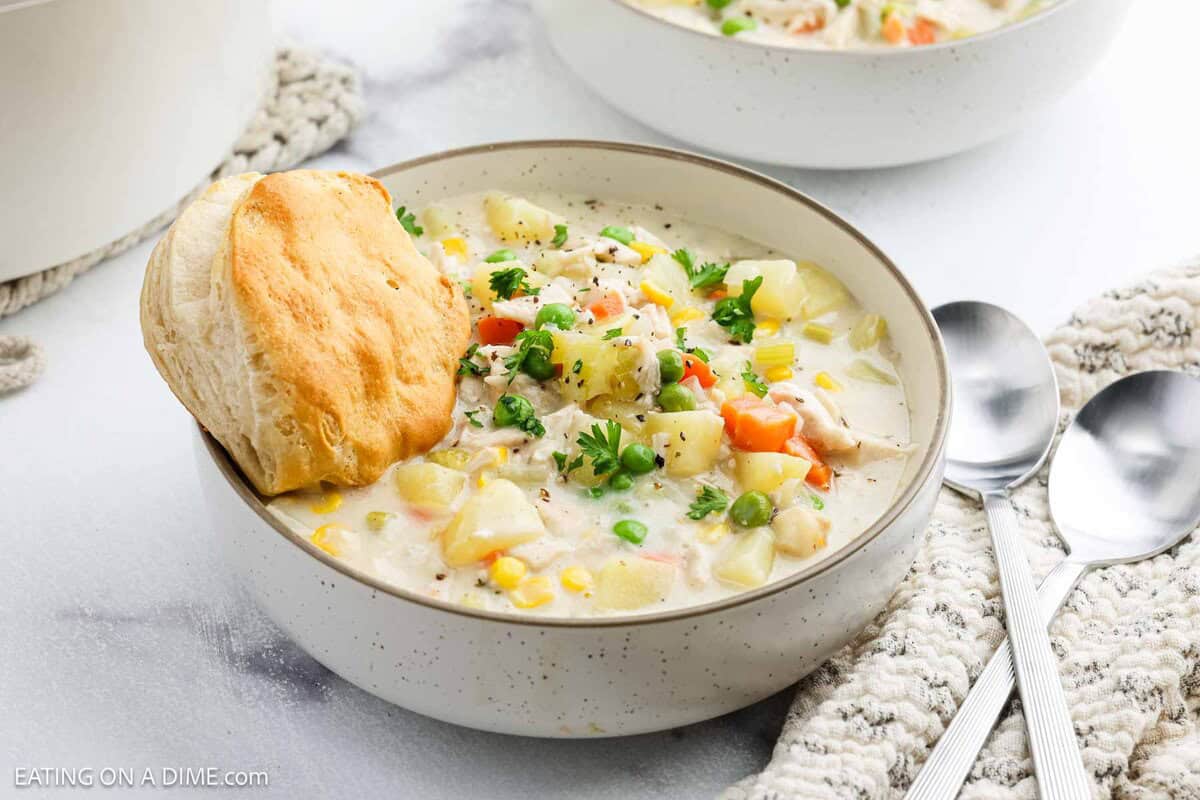 A bowl of creamy Chicken Pot Pie Soup, garnished with herbs and served with a golden biscuit on the rim. Spoons and a textured napkin lie beside the bowl on a white surface.