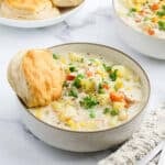 A bowl of creamy Chicken Pot Pie Soup with diced vegetables and herbs, served with a biscuit on the side. Another bowl of Chicken Pot Pie Soup and a plate of biscuits are in the background on a white surface.