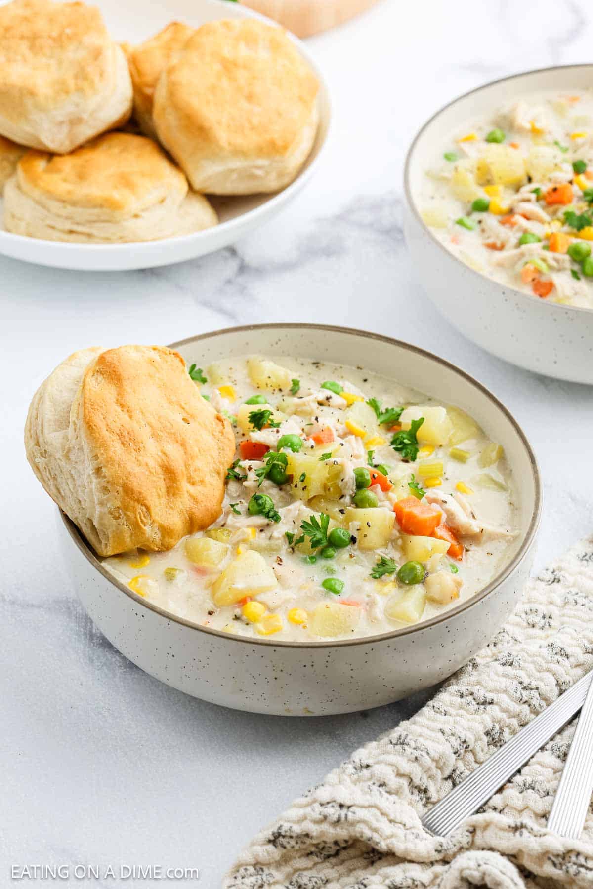 A bowl of creamy Chicken Pot Pie Soup with tender vegetables, garnished with parsley and served alongside a flaky biscuit. Another bowl of this comforting soup and a plate of biscuits sit invitingly in the background.