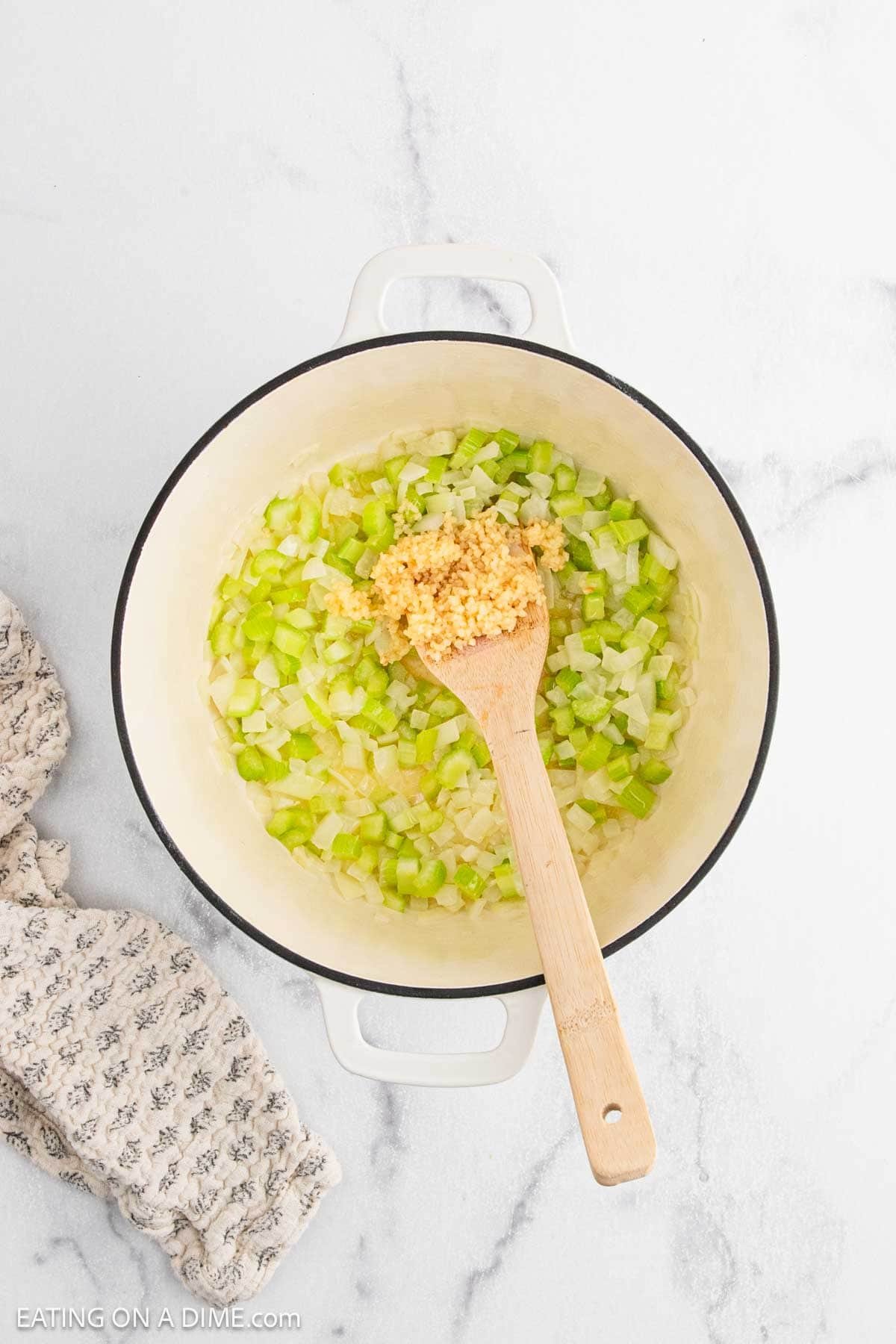A white pot with chopped onions and celery being sautéed for Chicken Pot Pie, a wooden spoon resting on top with minced garlic, on a marble countertop beside a patterned cloth.