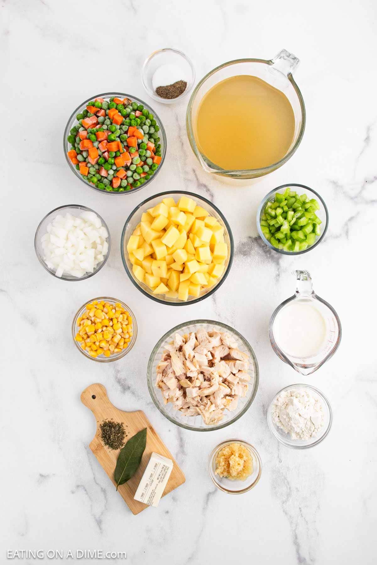 A flat lay of Chicken Pot Pie Soup ingredients in bowls: mixed vegetables, cubed potatoes, chicken broth, diced onions, peppers, corn, cooked chicken, milk, flour, garlic, butter, bay leaves, and pepper.