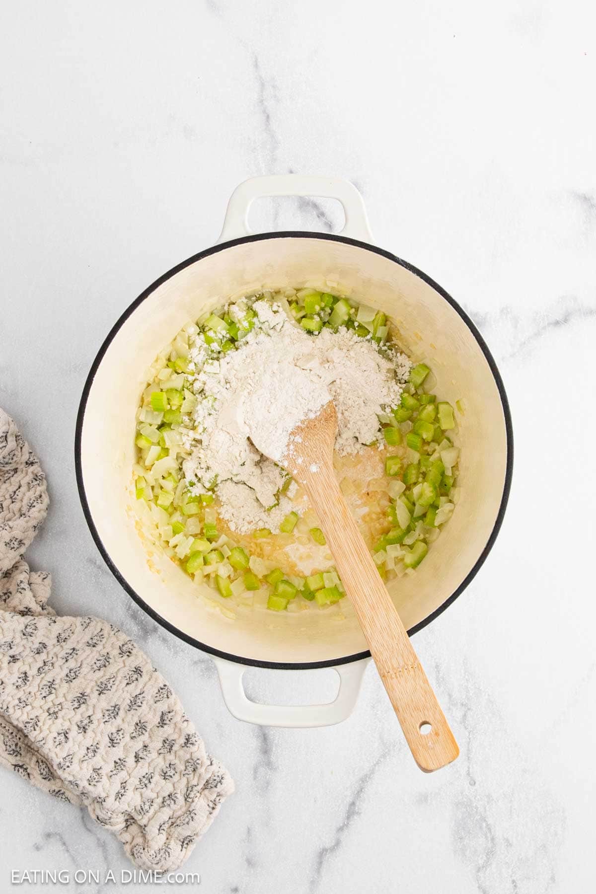A white pot filled with chopped celery and onions being sautéed for Chicken Pot Pie Soup, with a pile of flour on top and a wooden spoon resting inside. A patterned cloth napkin sits beside the pot on a white marble surface.
