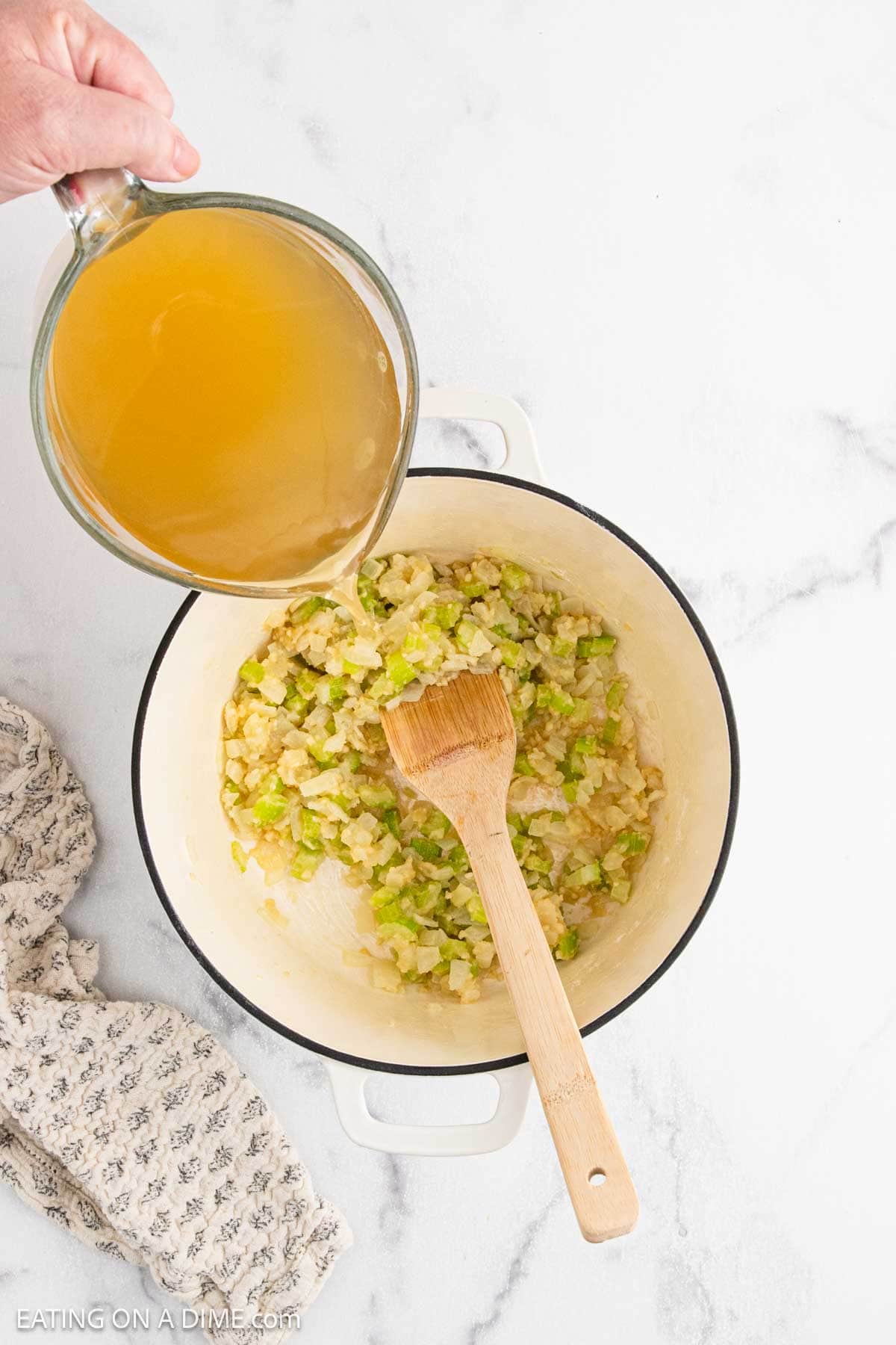 A hand pours broth from a glass measuring cup into a white pot of sautéed onions and celery, starting the base for Chicken Pot Pie Soup. A wooden spoon rests inside, and a patterned cloth sits beside the pot on a marble surface.