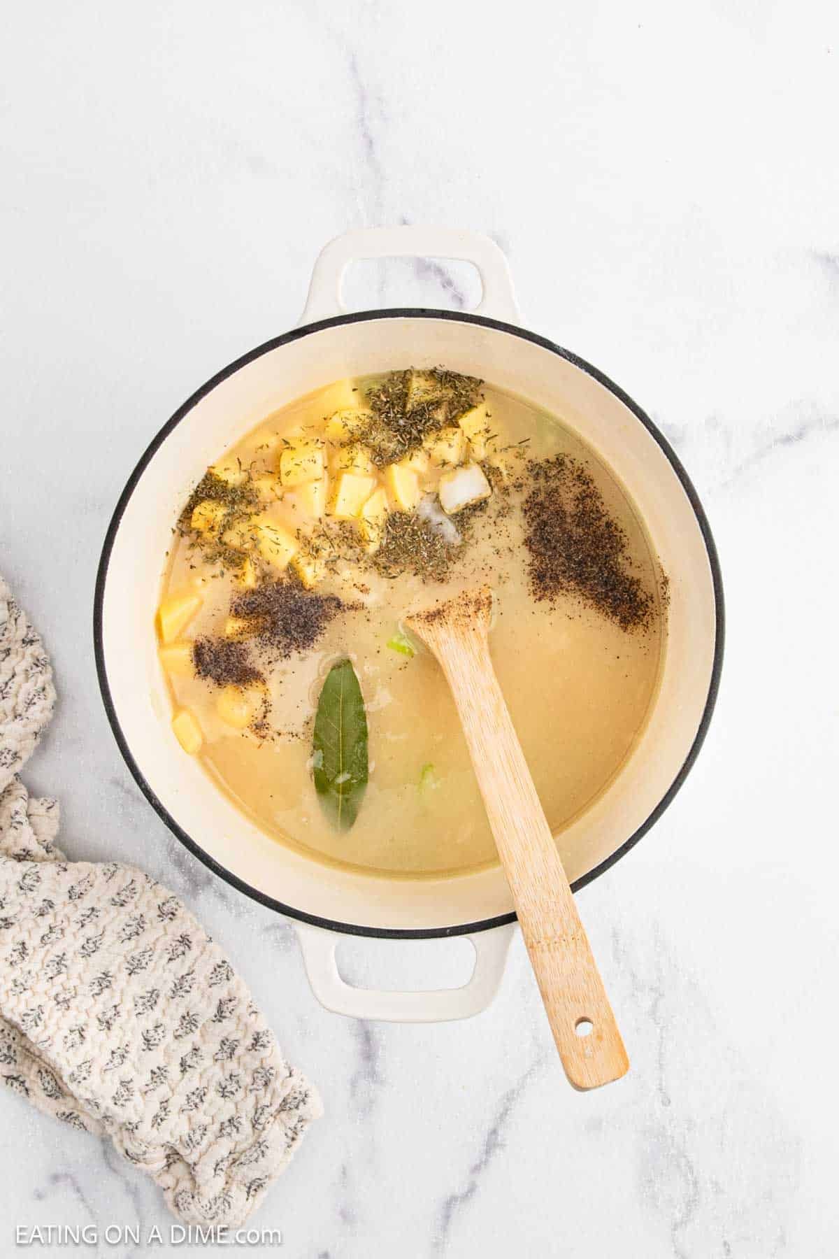 A white pot filled with broth, diced yellow cheese, seasonings, and bay leaves sits on a marble surface, ready to become a hearty chicken pot pie soup. A wooden spoon rests inside the pot, and a patterned cloth is nearby.