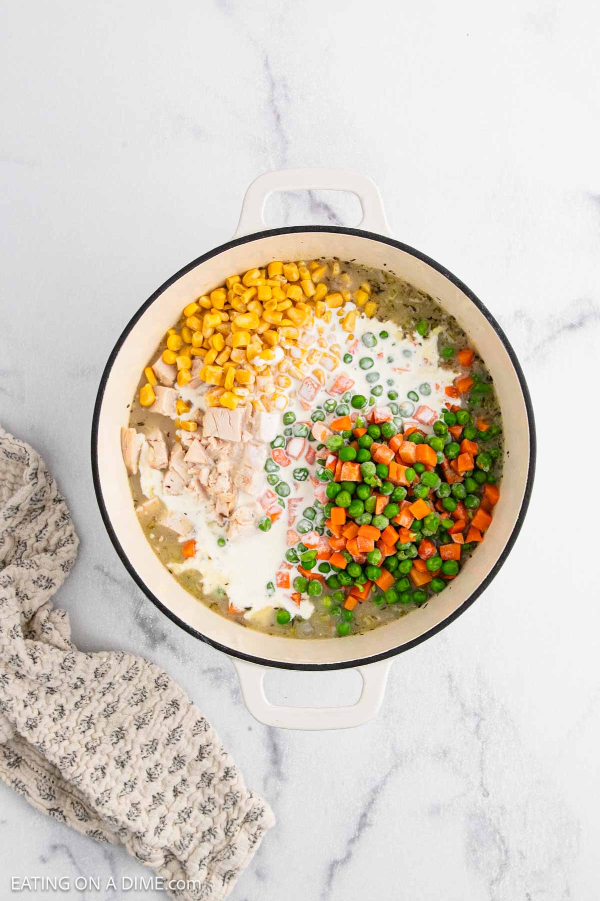 A white pot on a marble surface contains the ingredients for Chicken Pot Pie Soup—cream, corn, diced chicken, peas, carrots, and chopped green beans—all separated into sections before being mixed. A patterned towel lies to the left of the pot.