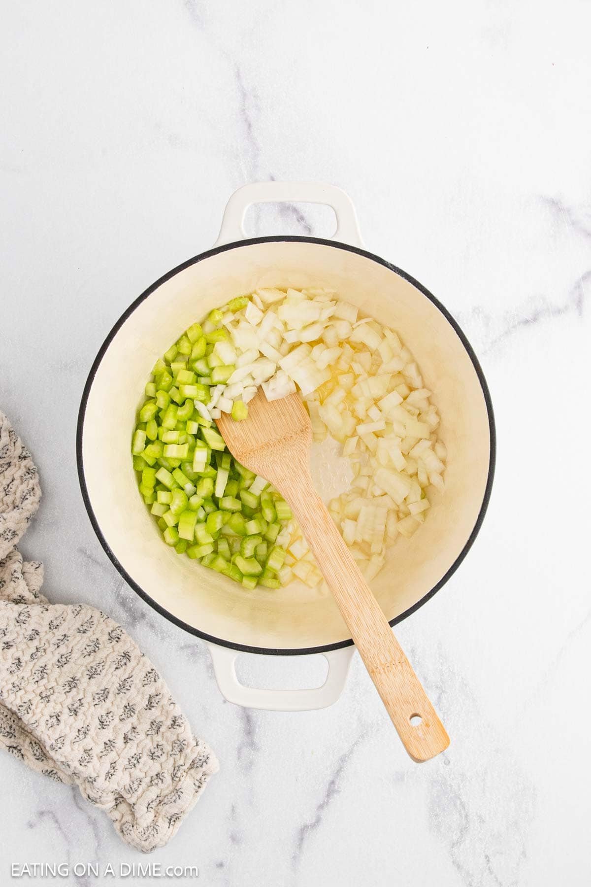 A white pot containing chopped onions and celery for Chicken Pot Pie sits on a marble surface with a wooden spoon resting inside. A patterned kitchen towel is placed beside the pot.