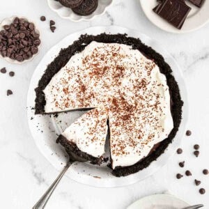 A decadent Chocolate Cream Pie with a dark cookie crust is topped with whipped cream and chocolate shavings. A slice is being served, surrounded by chocolate chips and pieces—a classic chocolate pie recipe on a white plate.