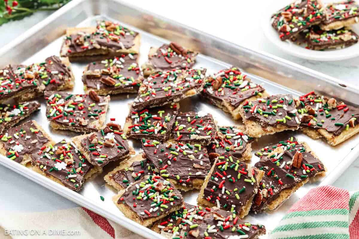 A baking tray filled with pieces of Christmas Crack toffee topped with chocolate and colorful red, green, and white sprinkles. A plate with more toffee pieces sits in the background, beside festive napkins.