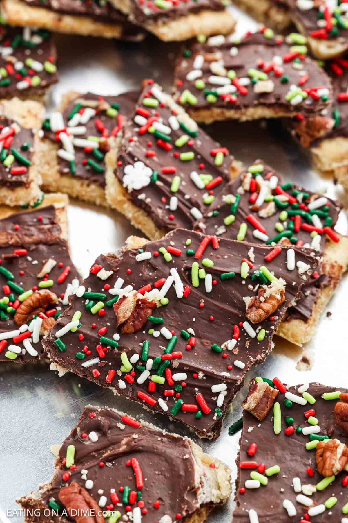 Pieces of Christmas Crack holiday toffee topped with chocolate, festive red, green, and white sprinkles, and chopped pecans are arranged on a flat surface.