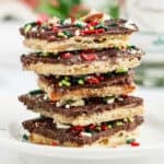 A stack of Christmas Crack chocolate-covered toffee crackers topped with festive red, green, and white sprinkles sits on a white plate.