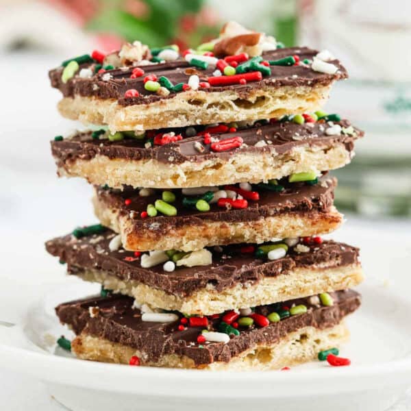 A stack of Christmas Crack chocolate-covered toffee crackers topped with festive red, green, and white sprinkles sits on a white plate.