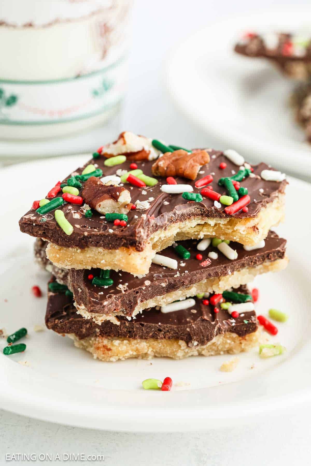 A stack of three homemade Christmas Crack chocolate toffee bark pieces topped with colorful red, white, and green sprinkles and pecan pieces sits on a white plate. A cup and more tasty Christmas Crack are visible in the background.