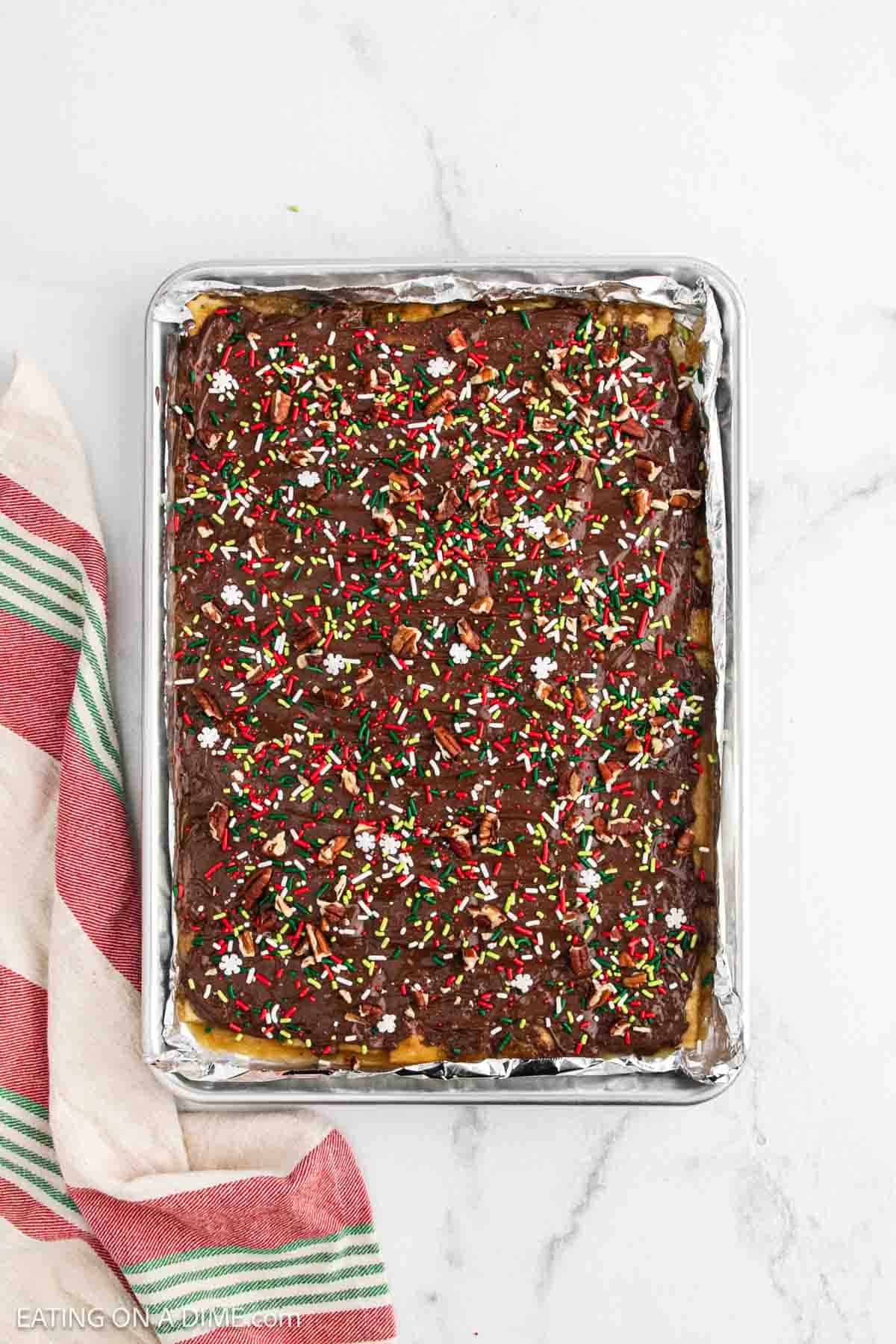 A baking sheet filled with Christmas Crack, a chocolate-covered dessert topped with red, white, and green sprinkles. The foil-lined sheet rests on white marble beside a red, green, and cream striped towel.