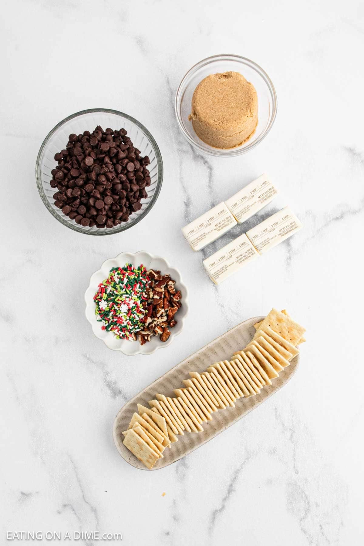 A flat lay of ingredients for Christmas Crack on a white surface: a bowl of chocolate chips, brown sugar, four butter sticks, colorful sprinkles, chopped pecans, and a plate of rectangular crackers.