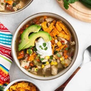 A bowl of chicken tortilla soup from Meal Plan 79 is topped with avocado slices, sour cream, fresh cilantro, and crunchy tortilla strips, surrounded by a colorful cloth and fresh ingredients.