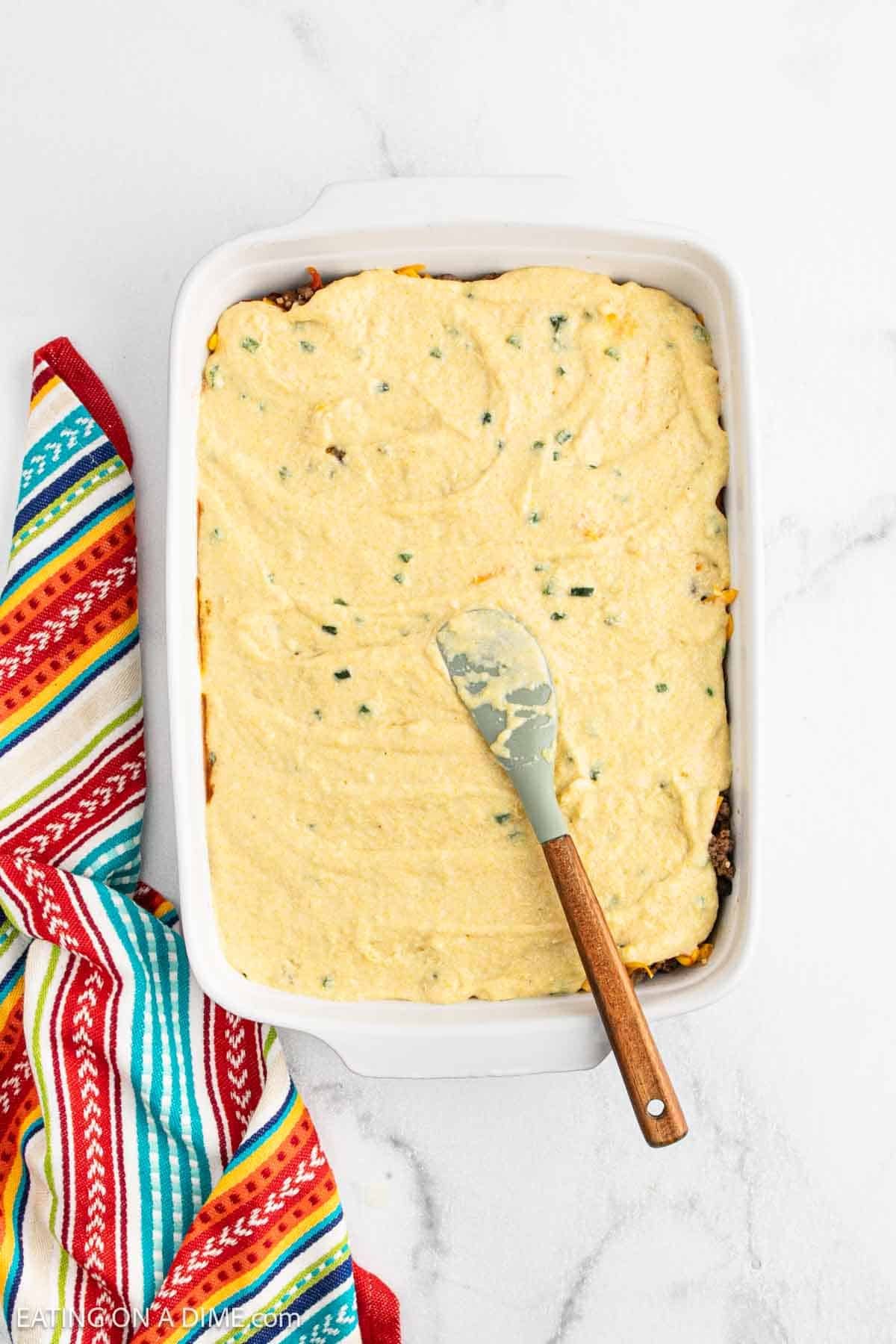 A baking dish filled with a Mexican Cornbread Casserole topped with a smooth, yellow cornbread layer, partially spread with a spatula. A colorful striped kitchen towel is beside the dish on a white countertop.