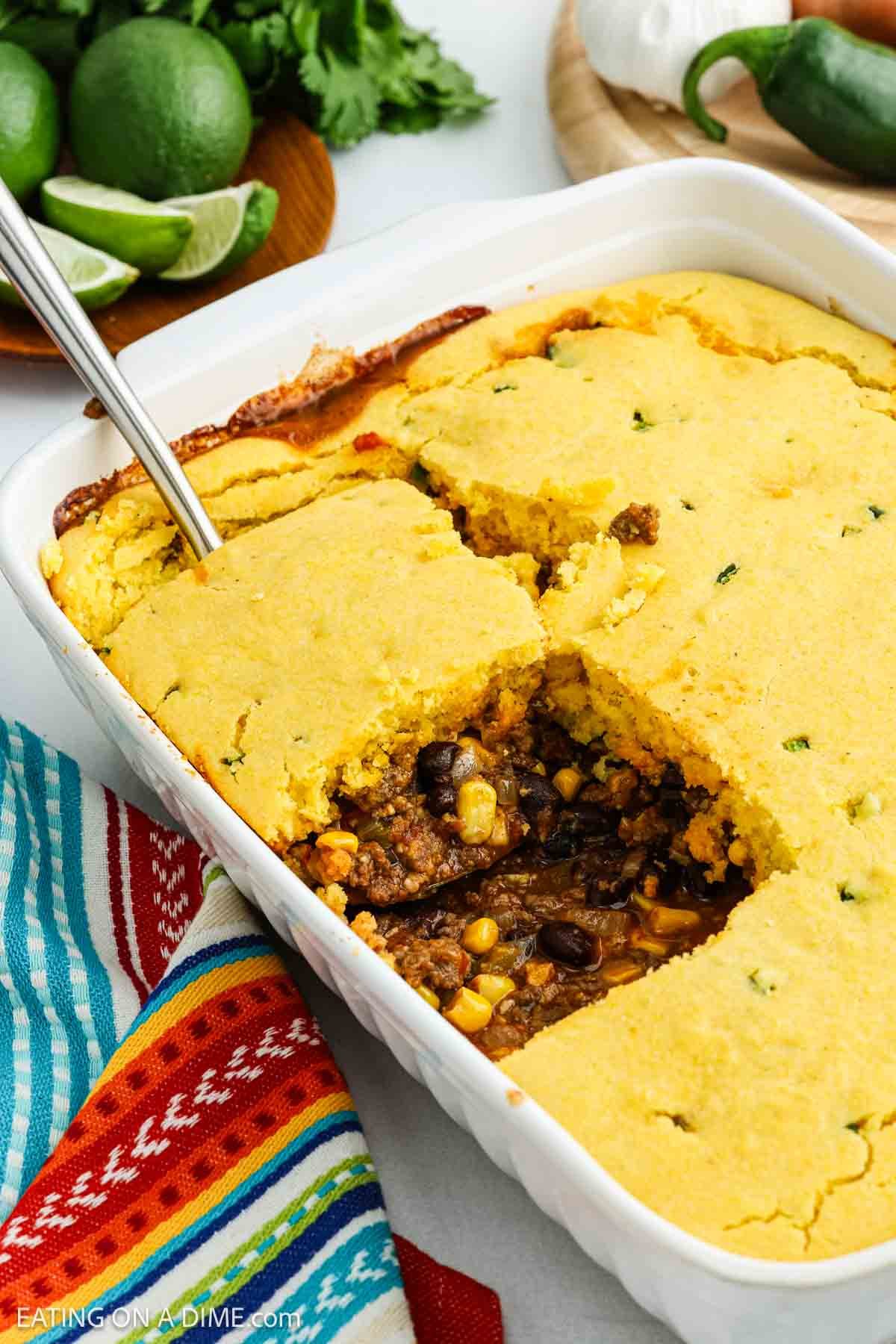 A casserole dish filled with Mexican cornbread casserole made of ground beef, black beans, and corn. A serving has been scooped out, revealing the filling. Limes, jalapeño, and a colorful towel are nearby.