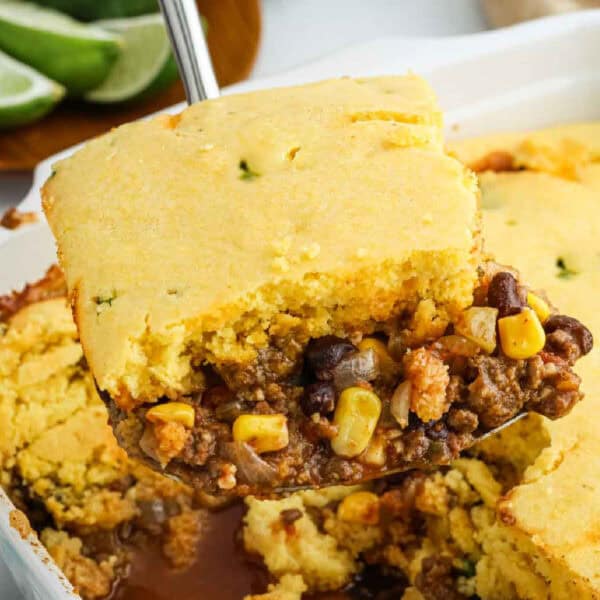 A serving of Mexican Cornbread Casserole with a golden cornbread topping, ground beef, black beans, corn, and onions is being lifted from a baking dish. Lime wedges are visible in the background.